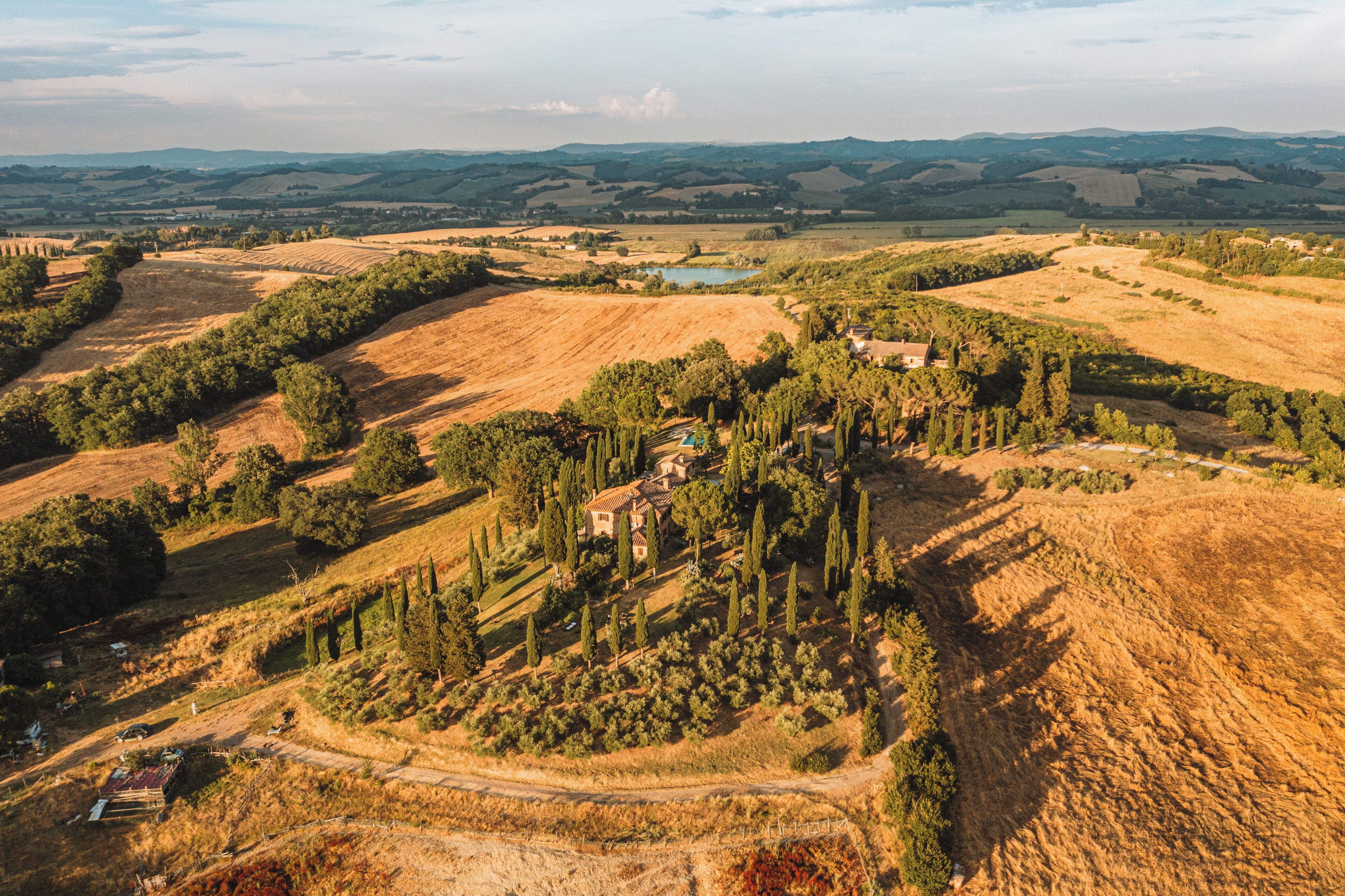 An aerial view of a rural landscape with rolling hills, a small cluster of buildings, and a body of water in the distance.