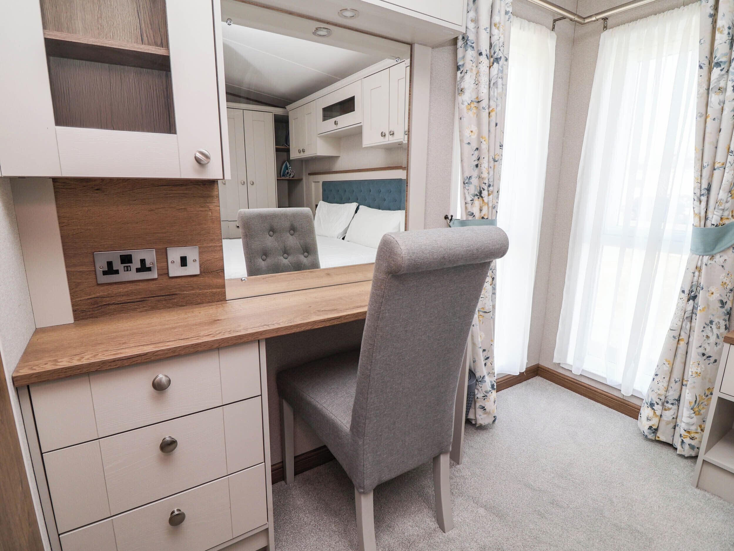 A compact room with a wooden desk, a grey upholstered chair, a mirror, and a bed with a blue headboard.