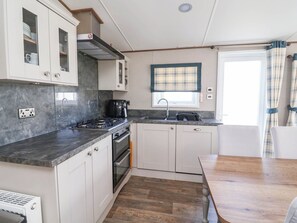 A compact kitchen with white cabinets, a gas stove, a coffee maker, and a window with a plaid curtain. - i35 - The Imperial (Abergele)