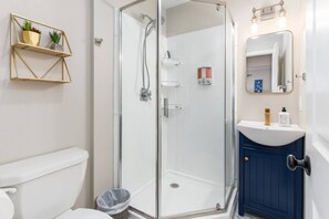 Ein Badezimmer mit einer Duschkabine aus Glas, einer weißen Toilette, einem blauen Schrank und einem an der Wand befestigten Regal mit Pflanzen.