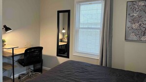 Ein Schlafzimmer mit Bett, Schreibtisch, Stuhl, Spiegel und einem Fenster mit Vorhängen.