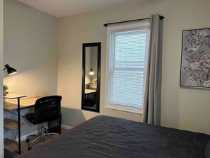 Ein Schlafzimmer mit Bett, Schreibtisch, Stuhl, Spiegel und einem Fenster mit Vorhängen.