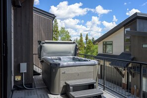 A hot tub on a balcony with a wooden deck, a metal railing, and a view of trees and a building in the background. - Luxury Emerald Escape | Hot Tub | Ski Shuttle (Winter Park)