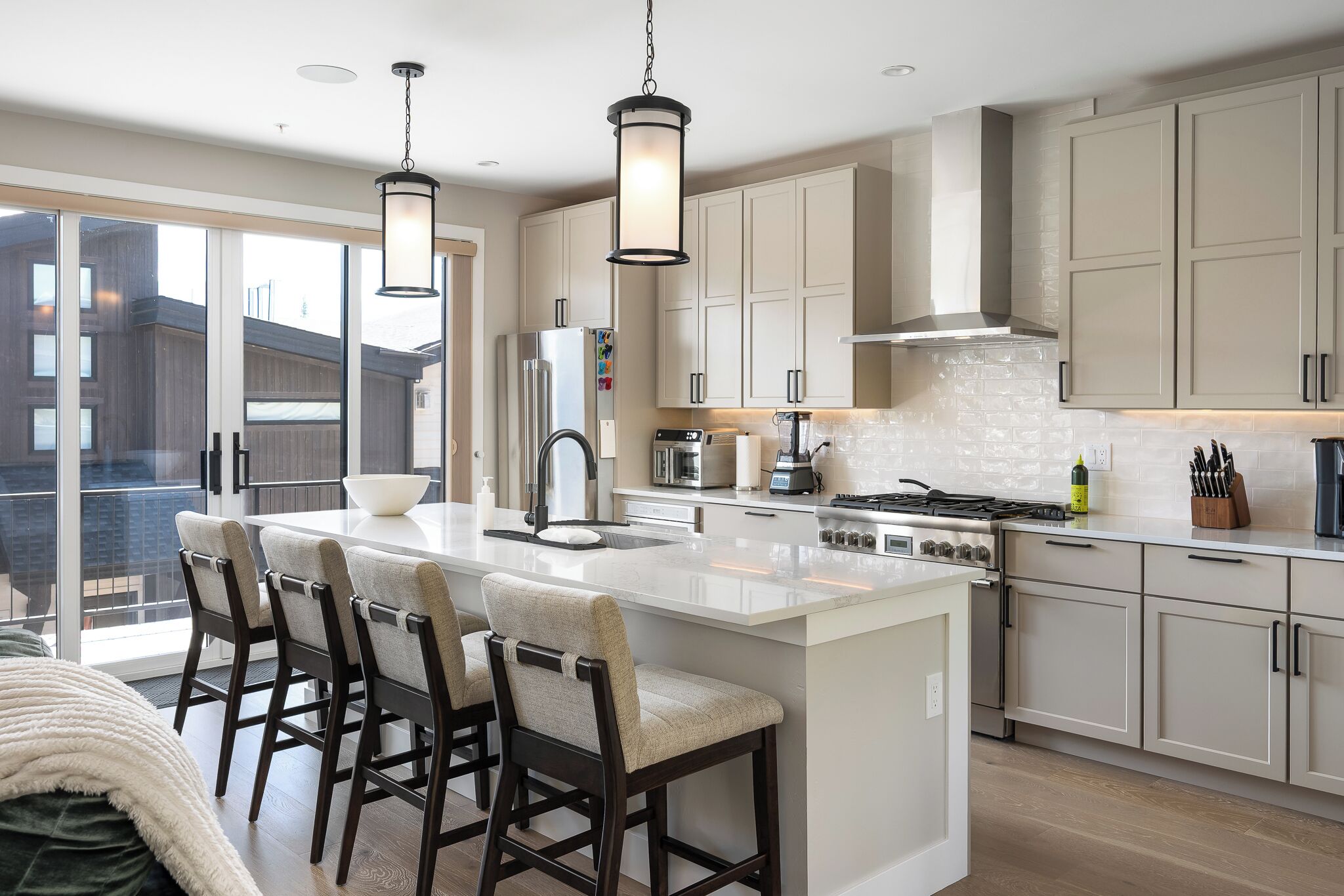 A modern kitchen with a central island, bar stools, a stainless steel stove, and a large window overlooking a balcony.