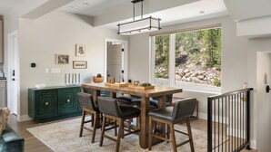 A dining area with a wooden table and chairs, a green cabinet, and a large window with a view of trees and rocks. - Luxury Emerald Escape | Hot Tub | Ski Shuttle (Winter Park)