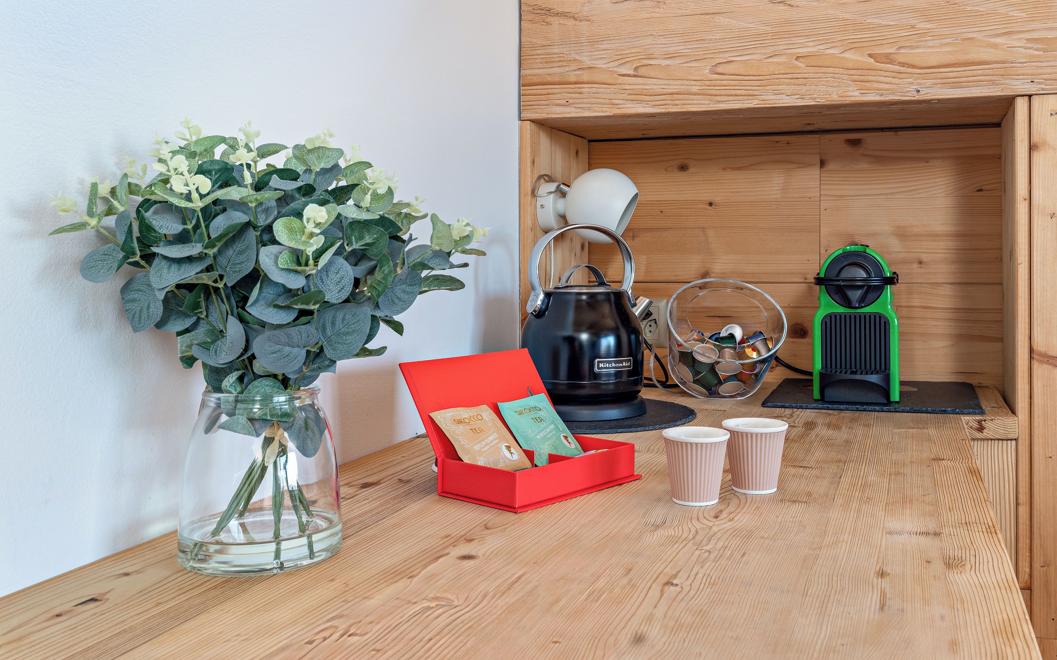 Una estantería de madera con un florero de vidrio con follaje verde, una tetera negra, una caja roja con bolsitas de té y una cafetera verde.