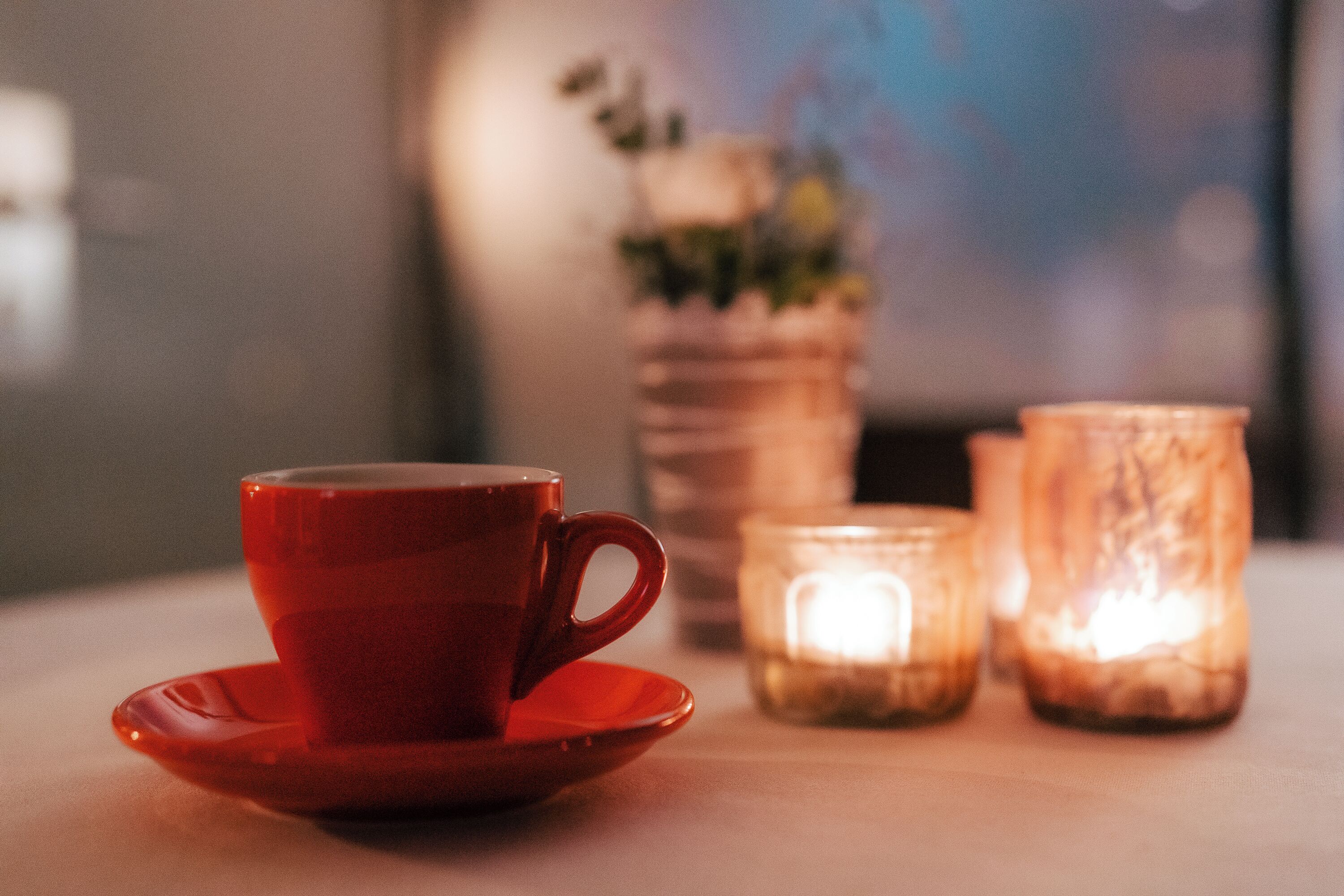 A red cup on a saucer, two lit candles, and a blurred vase with plants in the background.