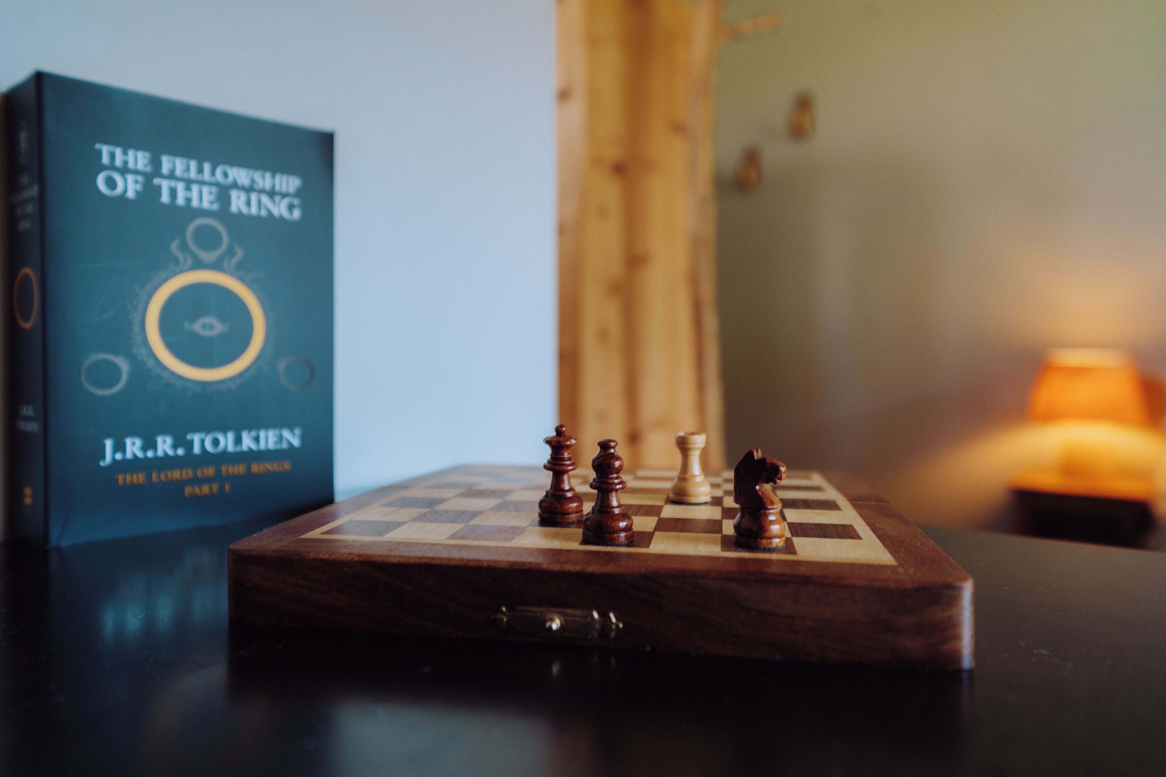 A chessboard with pieces, a book titled 'The Fellowship of the Ring' by J.R.R. Tolkien, and a lamp in the background.
