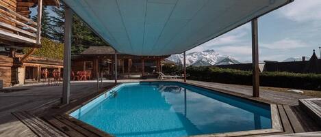 A swimming pool with a wooden deck, a covered seating area, and a mountain backdrop.