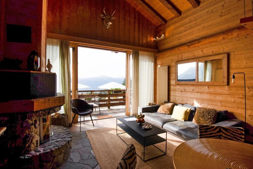 Un accogliente interno di una baita di legno con camino in pietra, un divano con cuscini, un tavolino da caffè e un'ampia finestra con vista sulle montagne.