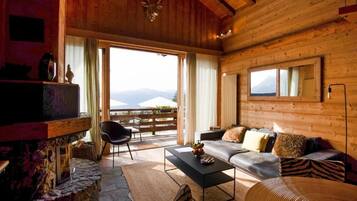 Ein gemütliches Holzhaus-Interieur mit Kamin aus Stein, einer Couch mit Kissen, einem Couchtisch und einem großen Fenster mit Blick auf die Berge.