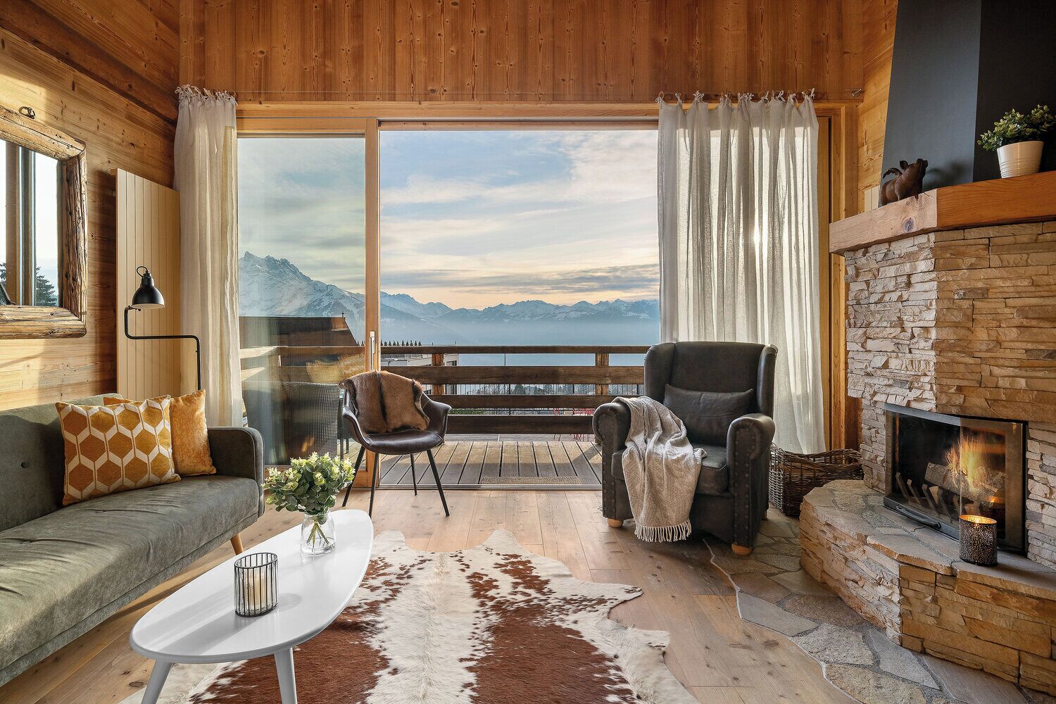 Una habitación acogedora con chimenea, un sofá, una silla y vistas a las montañas a través de amplias ventanas.