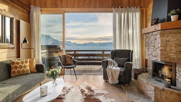 Una habitación acogedora con chimenea, un sofá, una silla y vistas a las montañas a través de amplias ventanas.