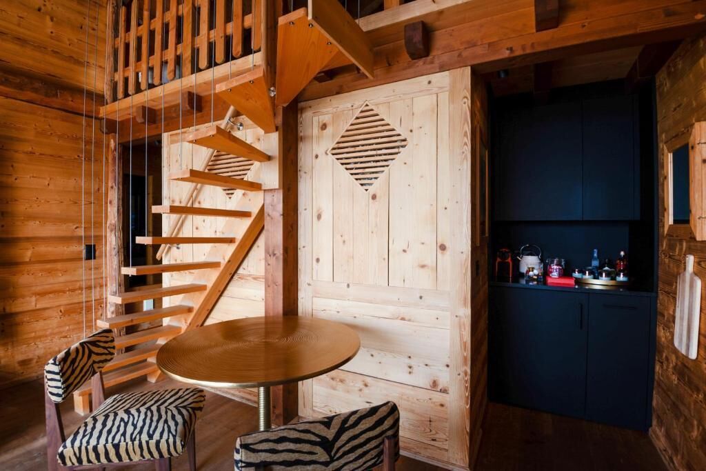 A wooden interior with a spiral staircase, a round table, zebra-patterned chairs, and a dark-colored cabinet.