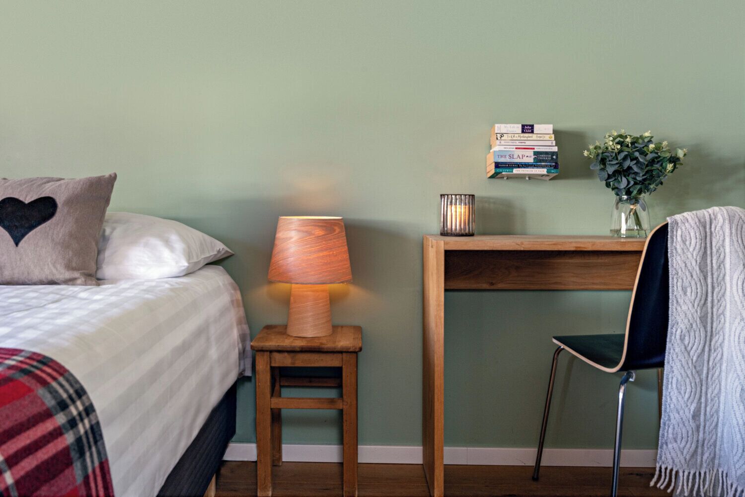 Ein Schlafzimmer mit einem Bett, einem Nachttisch mit Lampe, einem Sessel, einem kleinen Holztisch mit Büchern und einer Vase mit Blumen.
