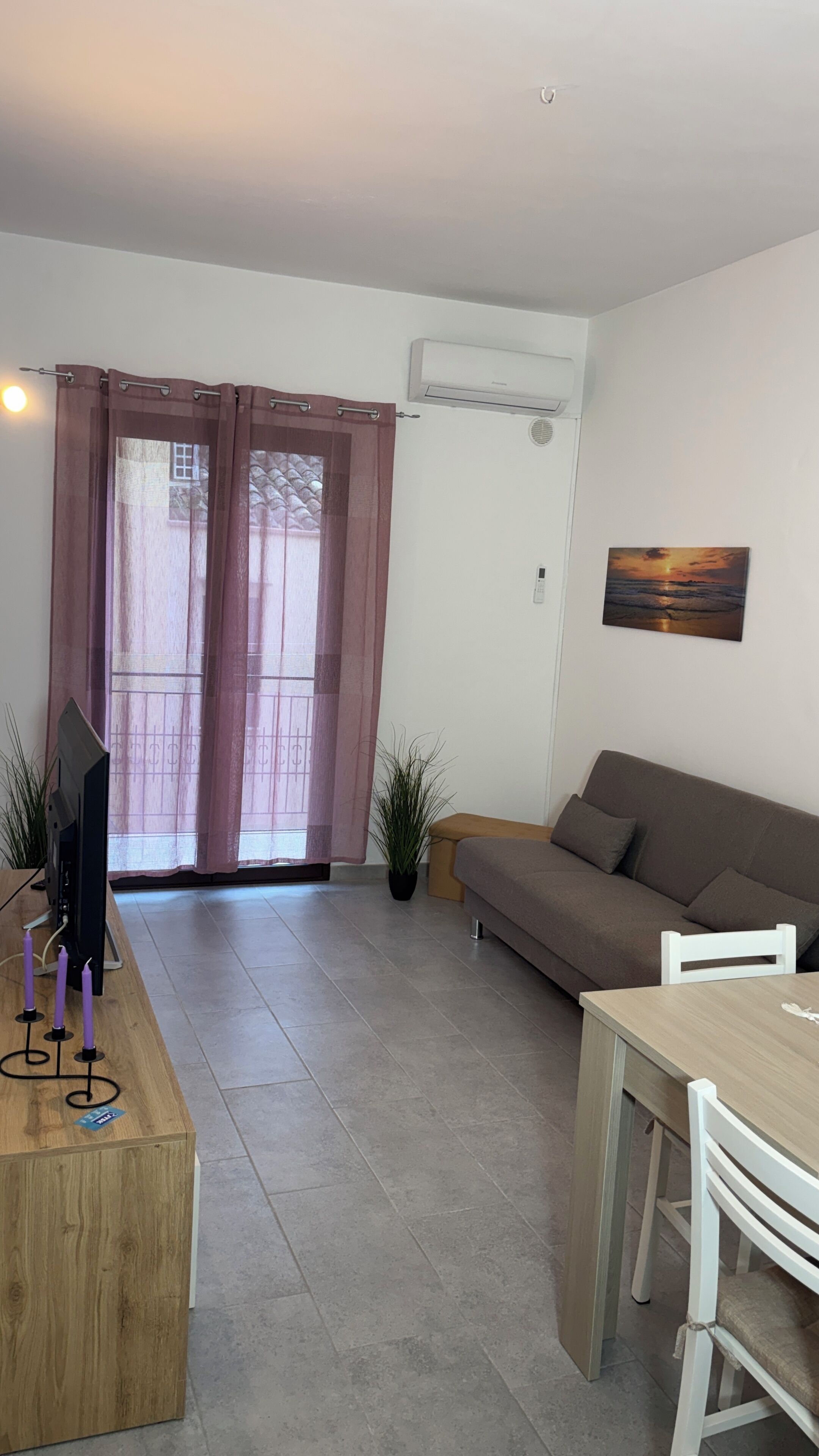 A living room with a sofa, a dining table, and a TV. There is a sliding glass door with curtains.