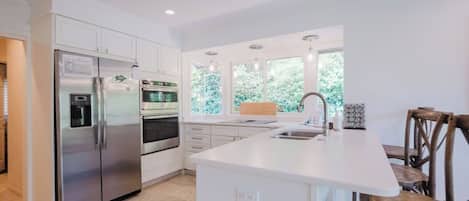 A modern kitchen with white cabinetry, stainless steel appliances, a central island, and a view of greenery outside.