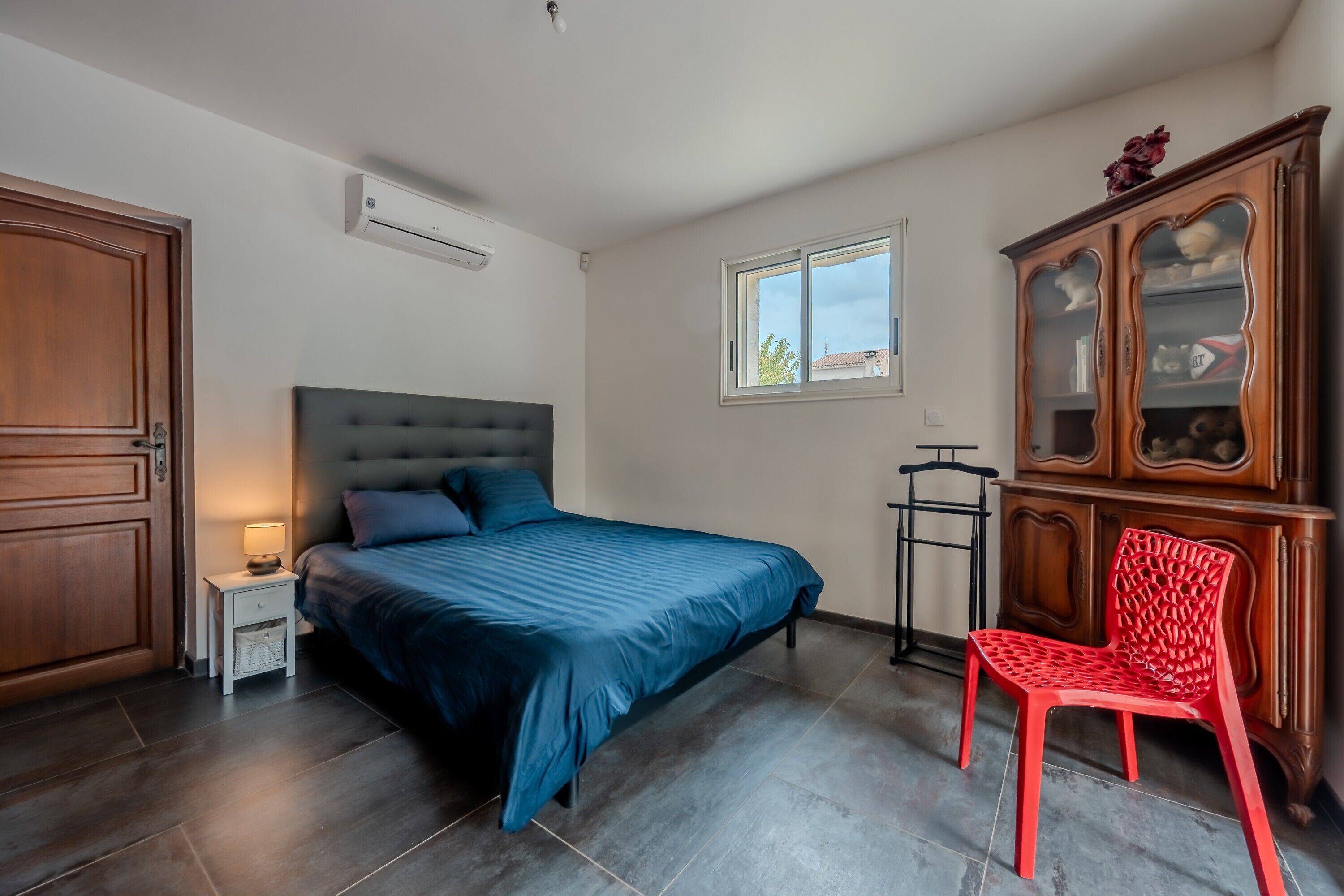 Ein Schlafzimmer mit einem Bett, einem Nachttisch, einem roten Stuhl, einem Holzschrank und einem Fenster.