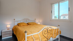 En seng med hvidt metalstel og et gult dynedække, et natbord med en lampe, og et vindue med udsigt til bygninger.