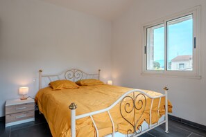 Uma cama com estrutura de metal branca e colcha amarela, uma mesinha de cabeceira com abajur e uma janela com vista para edifícios.