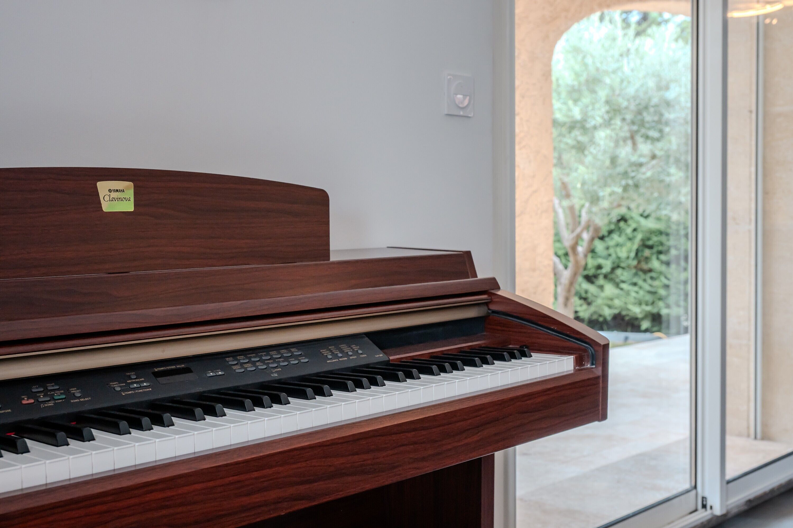 Ein Yamaha Clavinova Klavier mit Holzfurnier, platziert in einem Innenraum mit Blick auf Grünanlagen durch eine Glastür.