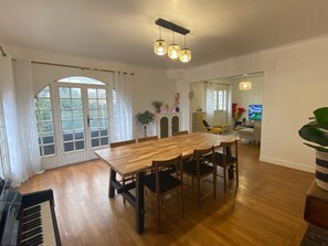 Een eetgedeelte met een houten tafel en stoelen, een piano en een groot raam met gordijnen.