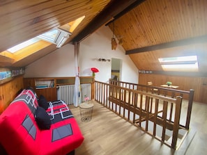 A cozy loft-style room with a wooden ceiling, a red sofa with geometric patterns, a small round table, and a view of a balcony area. - Lovely House with Splendid garden near Paris / Orly for 10-12 ppl (Épinay-sur-Orge)
