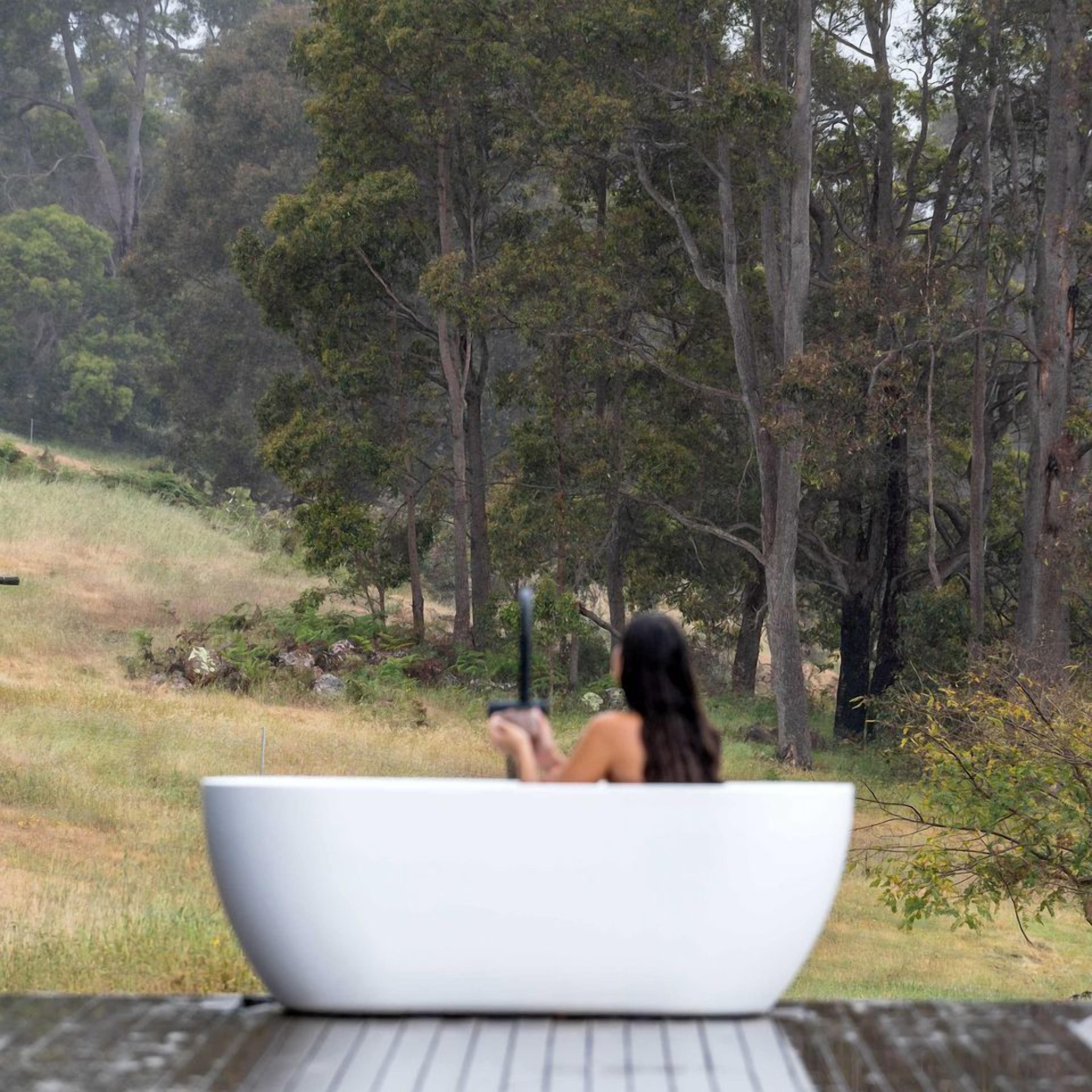 A person in a white bathtub outdoors with a natural landscape in the background.
