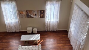 Um quarto com chão de madeira, um sofá branco, duas mesinhas redondas e uma janela grande com cortinas.