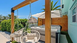 Una silla colgante de madera suspendida de una estructura de madera, un jacuzzi y un edificio con techo de paja en un jardín.