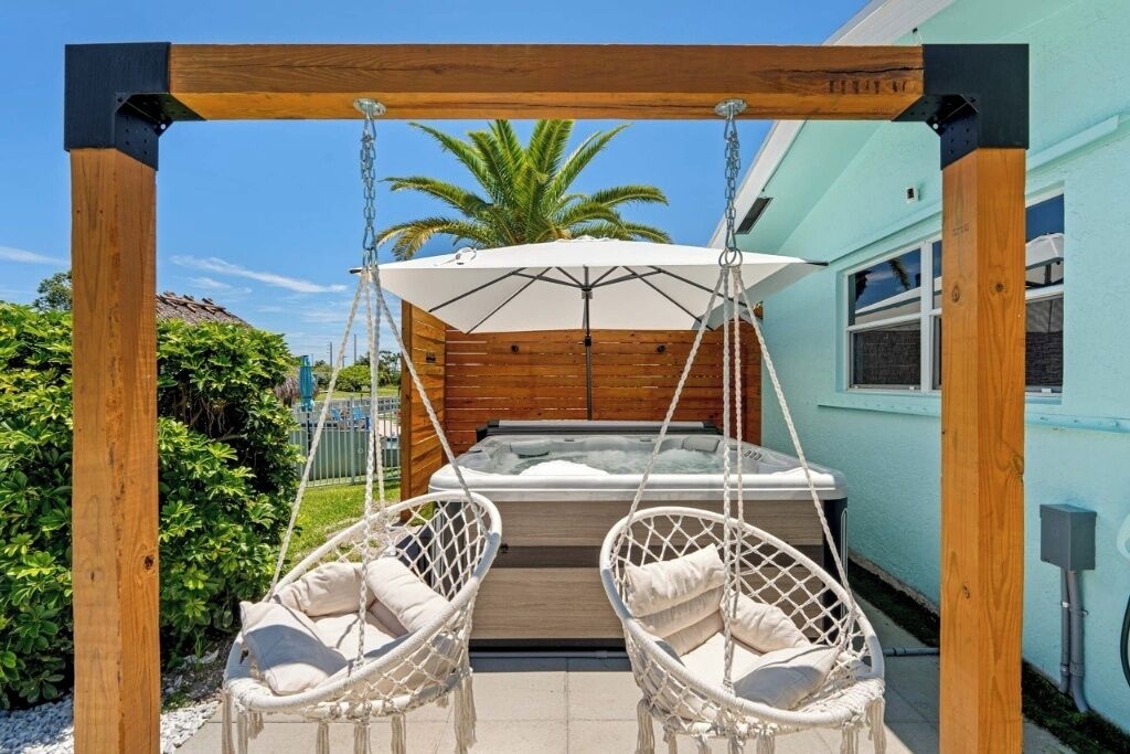 A wooden swing chair with a white cushion, a hanging white umbrella, and a hot tub in a backyard setting.