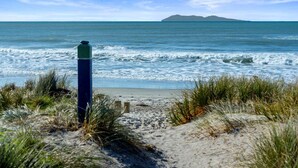 A beach with a grassy foreground, a sandy path leading to the water, and a distant island. - Bespoke Glamping Experiences in this Wonderful Rental in Wahihi Beach, New Zealand (Waihi Beach)