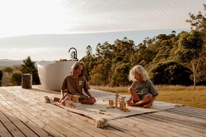 Two individuals sitting on a wooden deck outdoors with a large white bathtub and wooden blocks in the foreground.