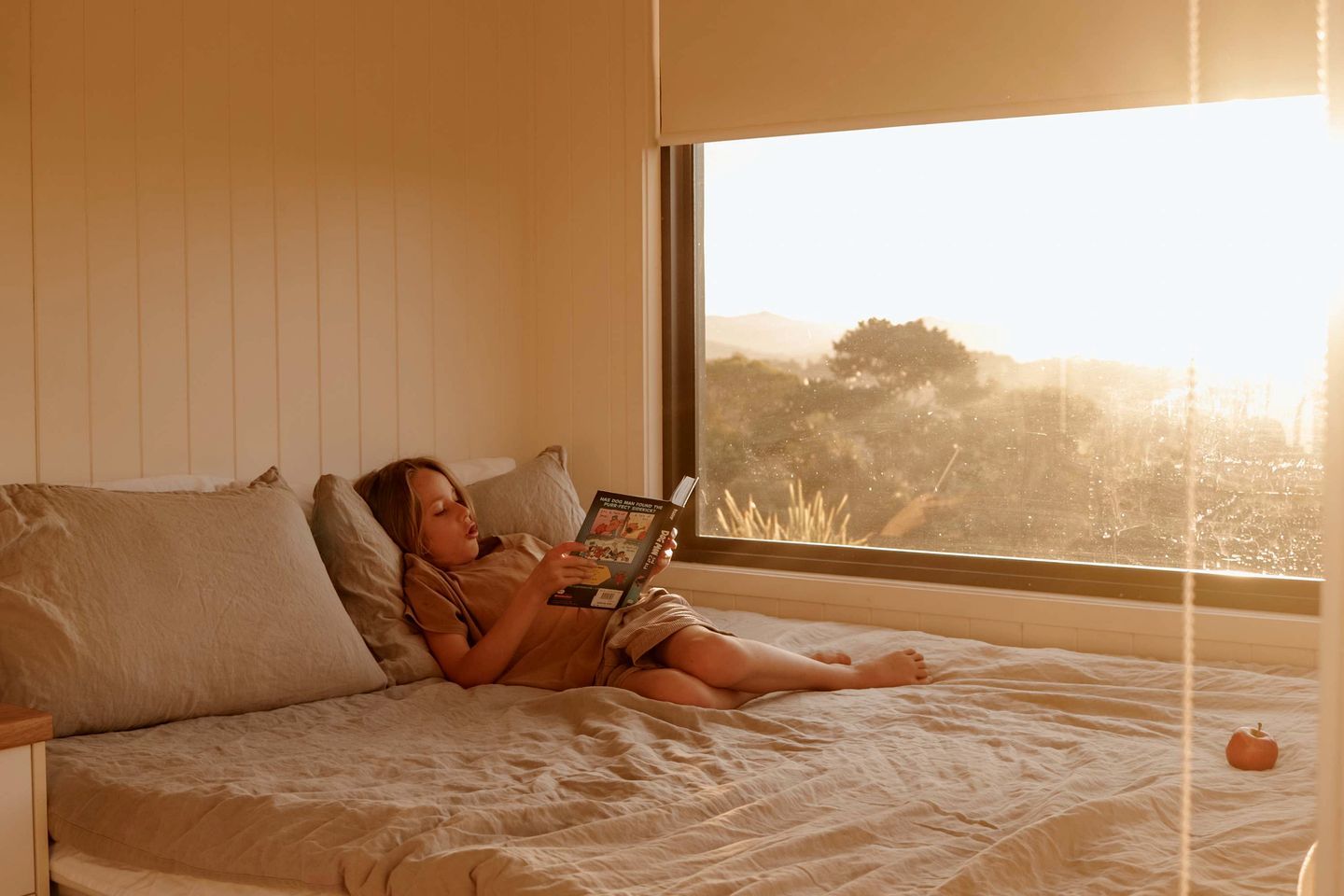 Une personne allongée sur un lit, un livre à la main, dans une pièce avec une fenêtre donnant sur des arbres.