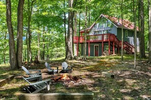 A cabin with a red porch, surrounded by trees, and a fire pit with chairs.