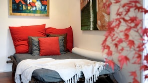 Ein Bett mit roter und grauer Bettwäsche, einer weißen Tagesdecke und einem Bild eines Hauses an der Wand.