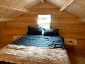 A wooden cabin bedroom with a bed, pillows, a window, and a nightstand.