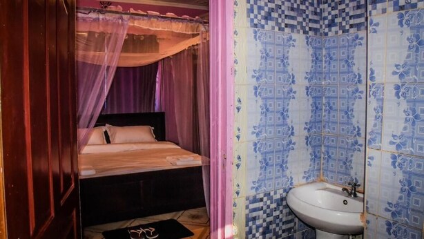 Un dormitorio con cama, mesita de noche, ventana con cortina y baño con lavamanos y paredes de azulejos.