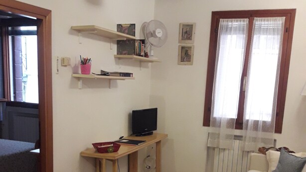 Um quarto com piso de madeira, uma mesa com computador, uma cadeira, uma cama e uma janela com cortinas.