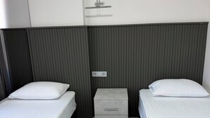 Ein Hotelzimmer mit zwei Einzelbetten, einem kleinen Nachttisch, einer Kommode und einem an der Wand befestigten Regal.