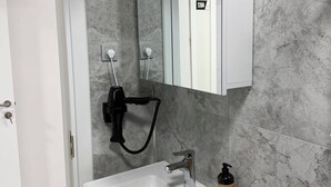 Ein Badezimmer mit einem weißen Waschbecken, einem Spiegel und einem Fön auf einem an der Wand befestigten Stativ.
