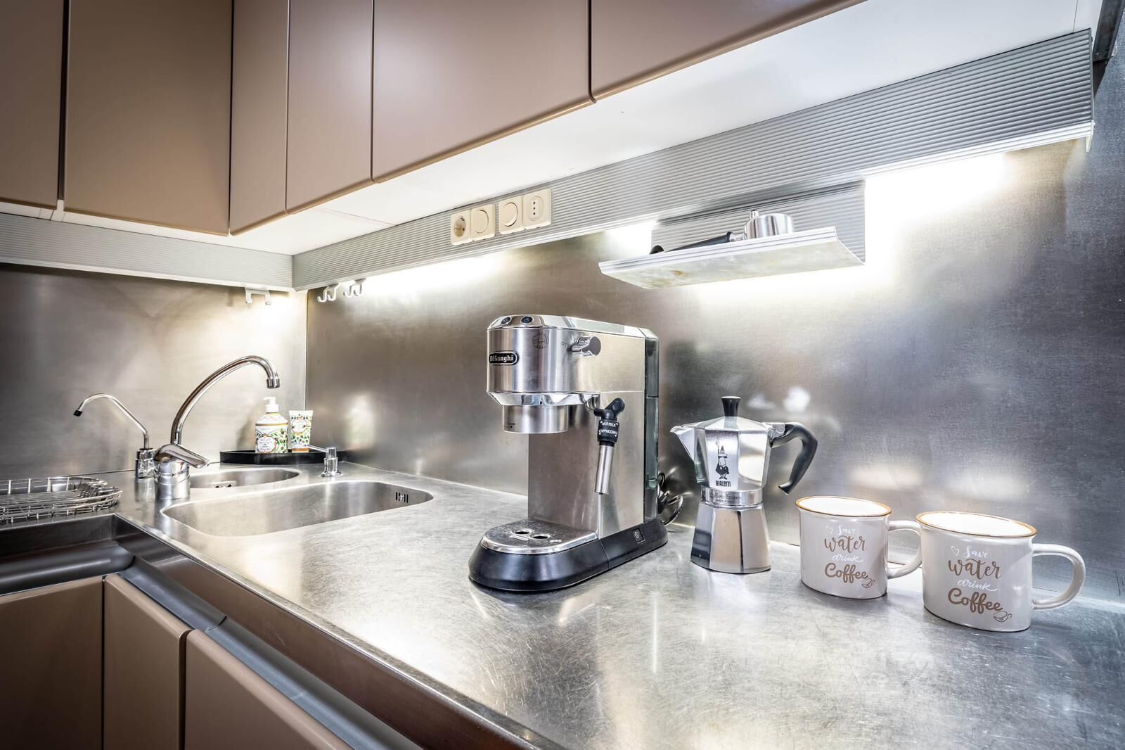 A modern kitchen with stainless steel appliances, including a coffee maker and a kettle, and two mugs with coffee-related text.