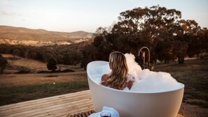 Une personne se détend dans une baignoire remplie de mousse, avec une vue panoramique sur les collines et les arbres en arrière-plan.