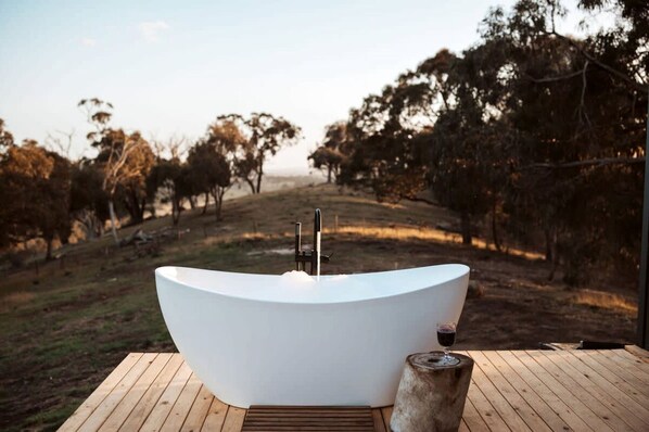 A large, white freestanding bathtub on a wooden deck with a wine glass on a stone pedestal, overlooking a vineyard.