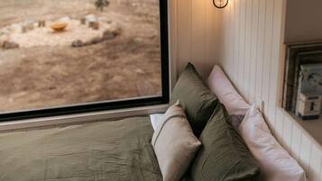 Ein Zimmer mit Bett, Kissen, Nachttisch, Fenster mit Blick auf einen Außenbereich und einer Wandleuchte.