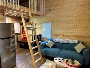 A wooden interior with a blue sofa, a ladder, a refrigerator, and a window with blinds.