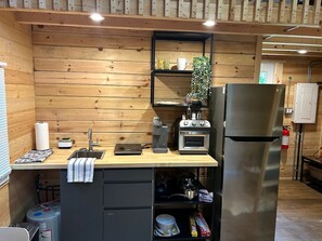 A compact kitchen with wooden walls, a sink, a coffee machine, a toaster, a refrigerator, and a shelf with kitchen items.