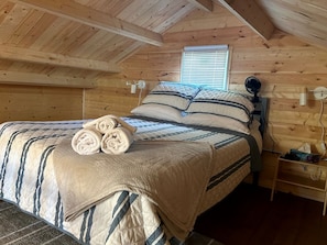 A wooden-paneled room with a bed, a bedside table, a fan, and a window.