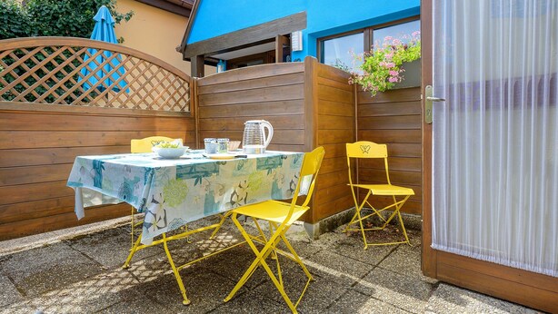 Un patio al aire libre con una mesa dispuesta para dos, sillas amarillas, una valla de madera y un edificio azul al fondo.