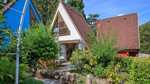 Una casa con techo inclinado, puerta principal visible y jardín con diversas plantas y un pequeño arroyo.
